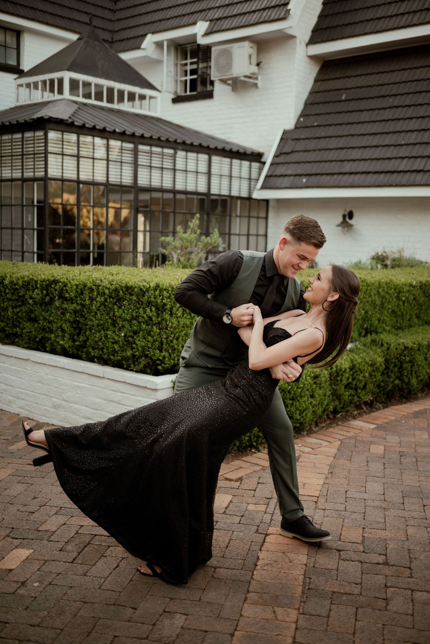 Couple dancing romantically on brick patio outside white house with black trim and manicured hedges.