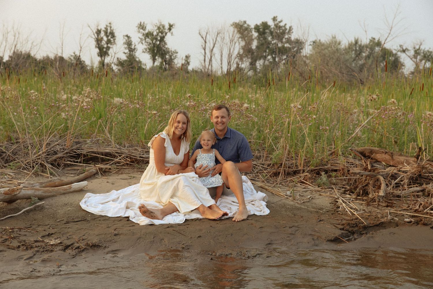 Couple in casual attire posing together on beach with driftwood and grass.