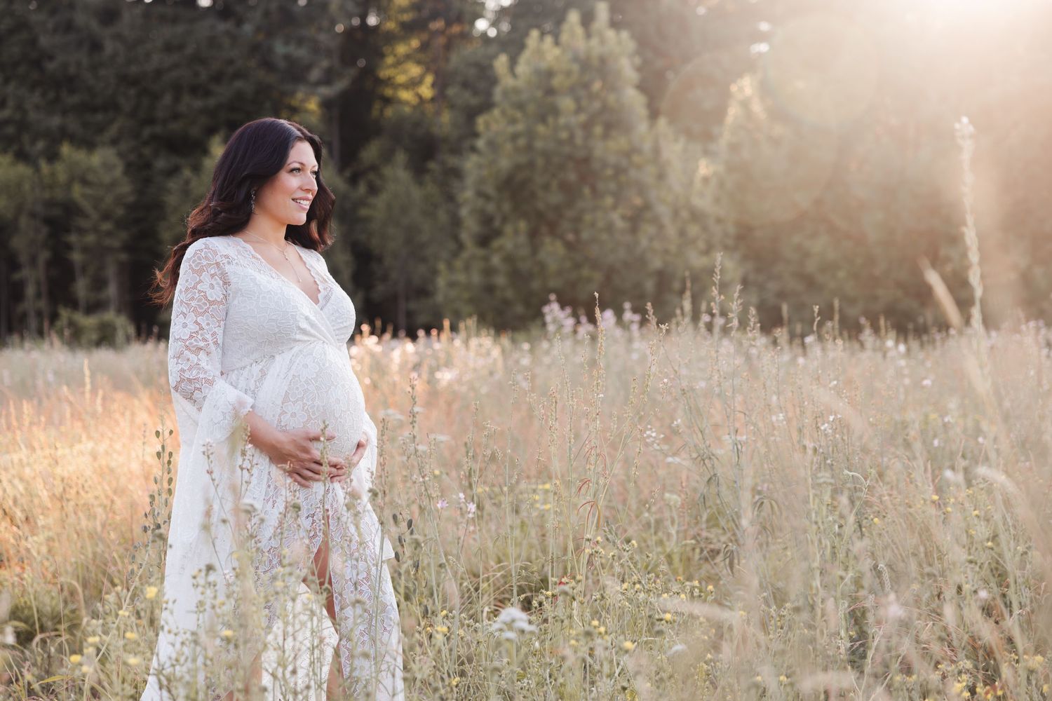A pregnant woman in a white lace dress stands in a sunlit meadow during golden hour.