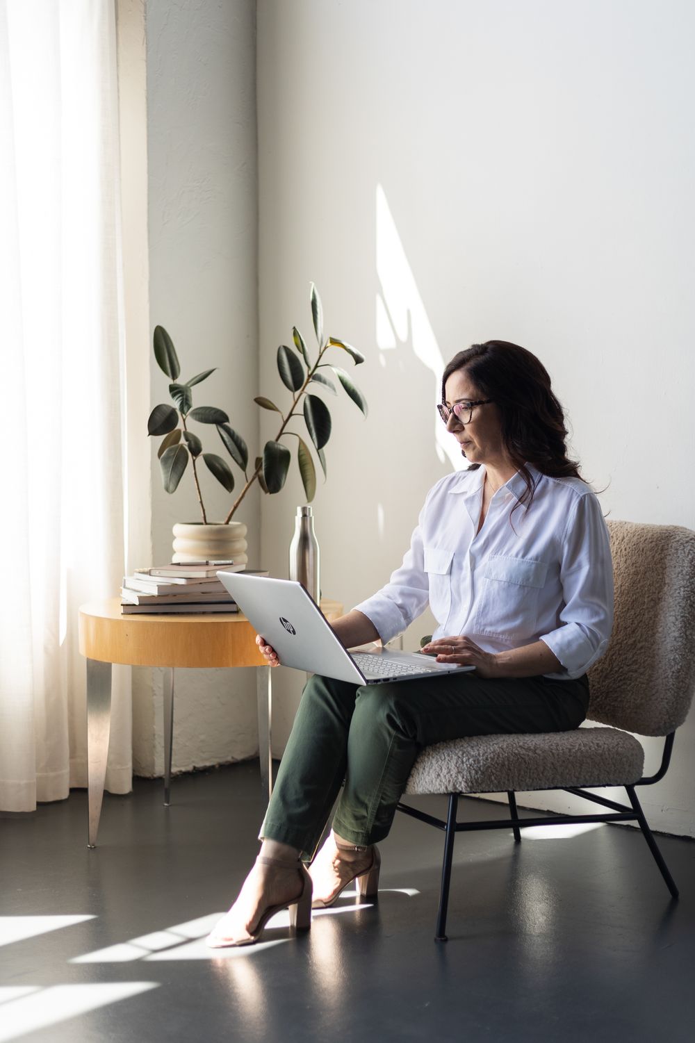 A person in a white shirt and green pants works on a laptop while sitting in a modern gray chair near houseplants.