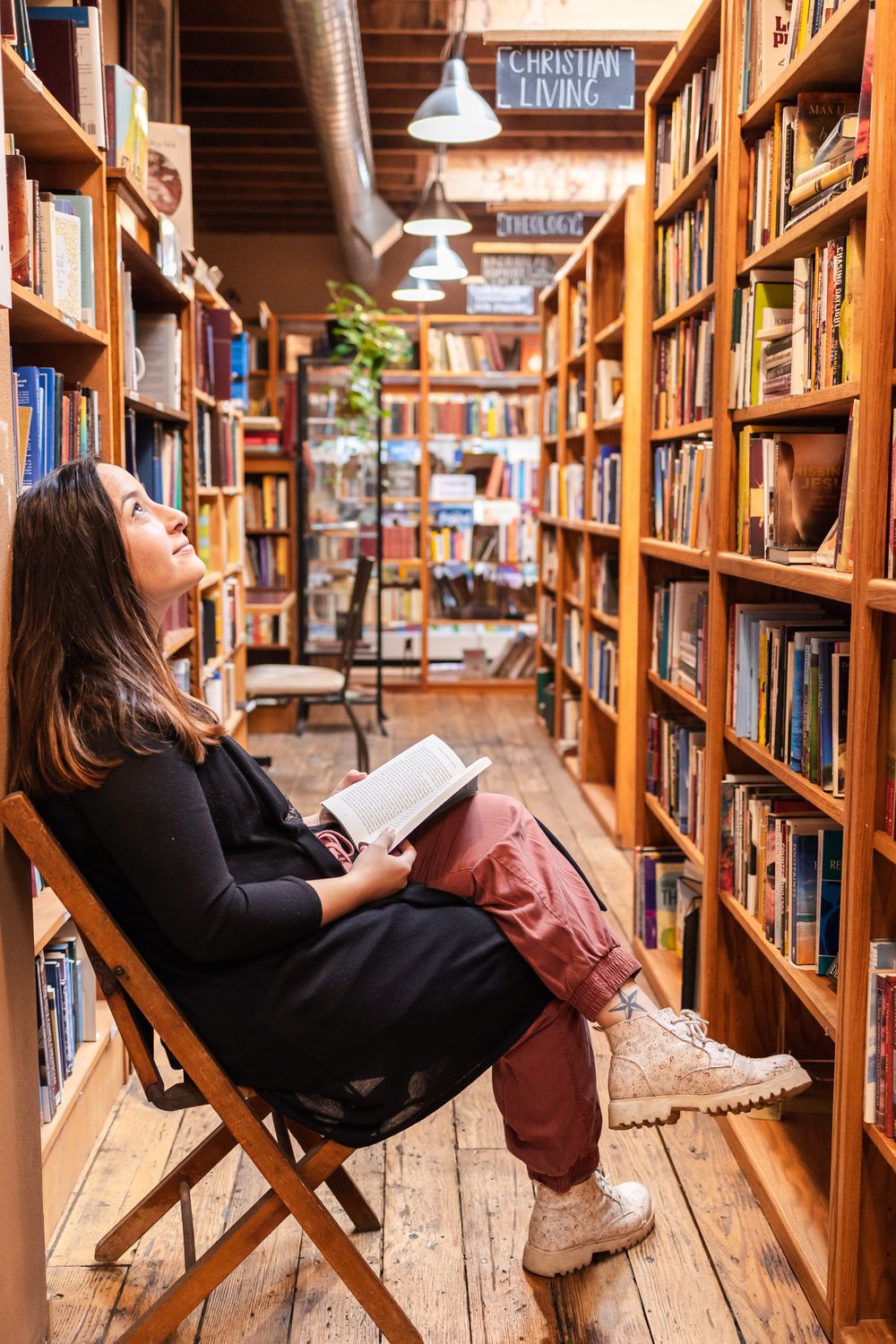 A person relaxes in a wooden chair while reading between bookshelves at a cozy bookstore with warm lighting.