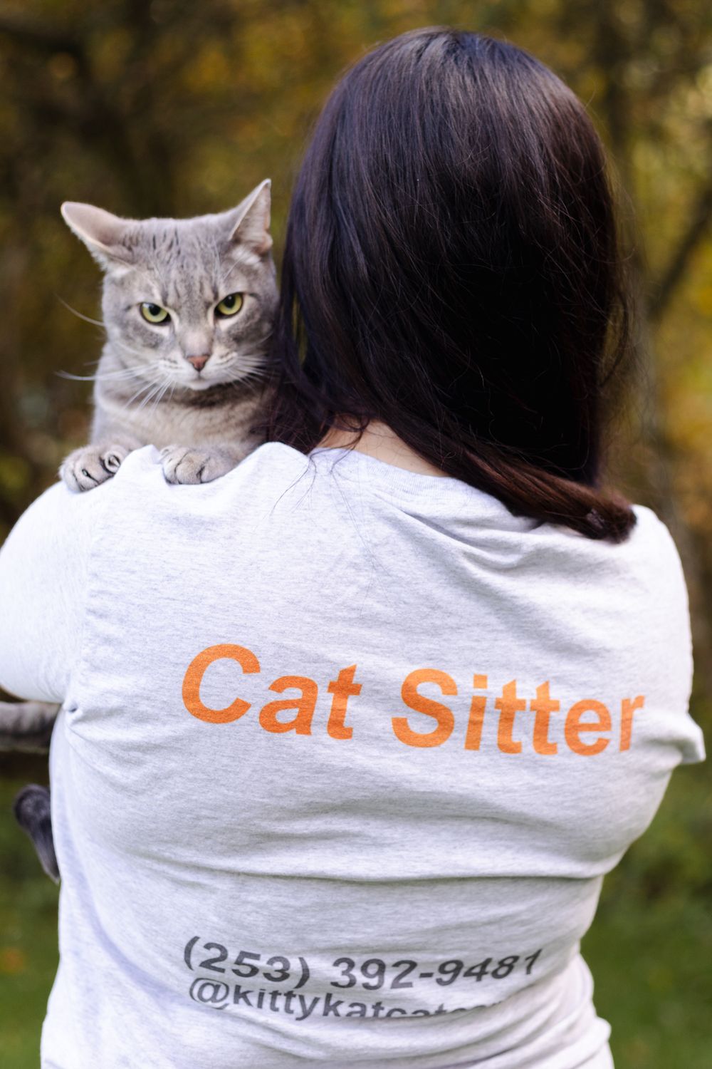 A cat peeks over someone's shoulder wearing a white t-shirt with 'Cat Sitter' text and phone number printed on the back.