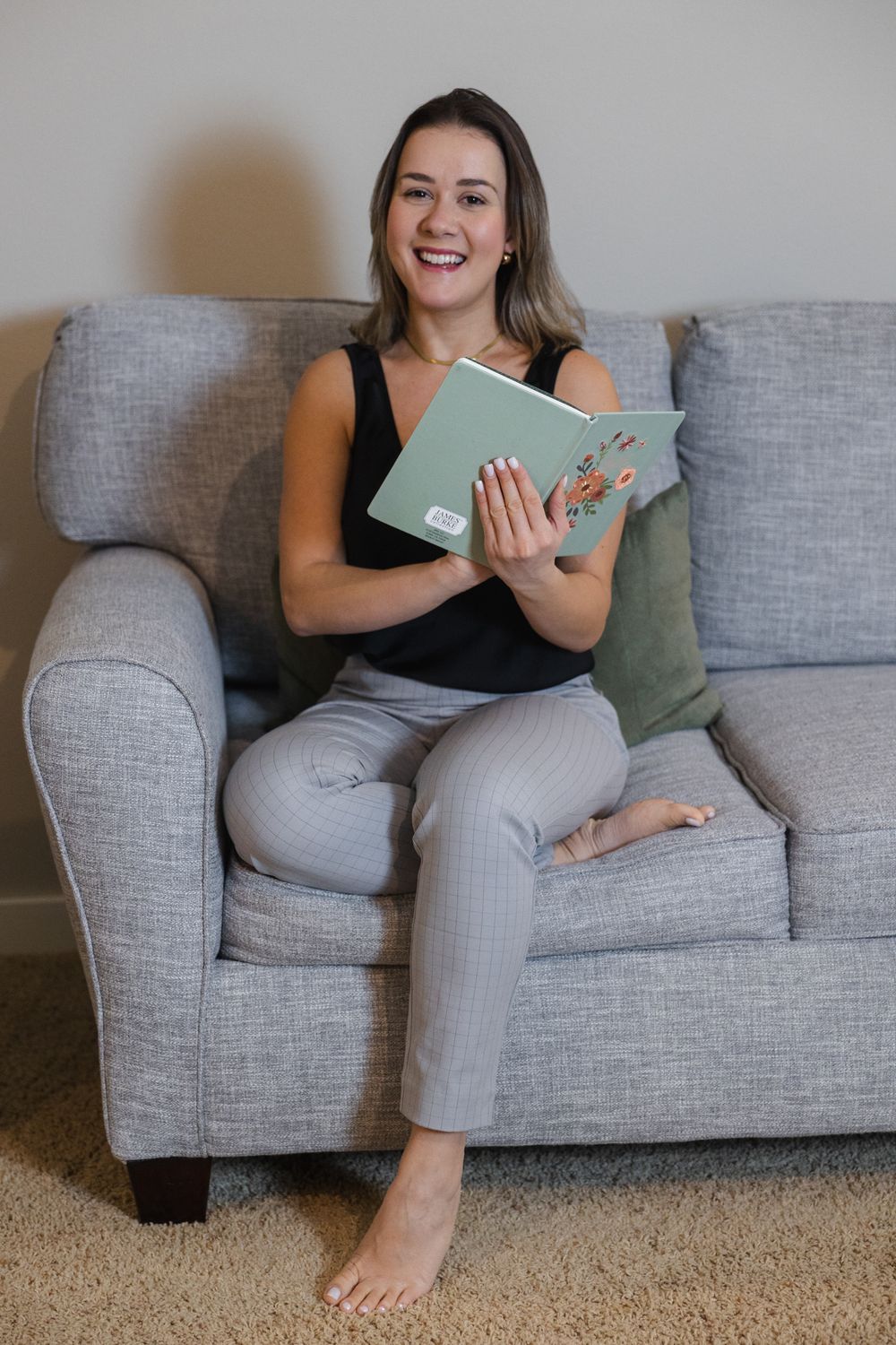 Someone lounges comfortably on a gray couch while reading a book and smiling.