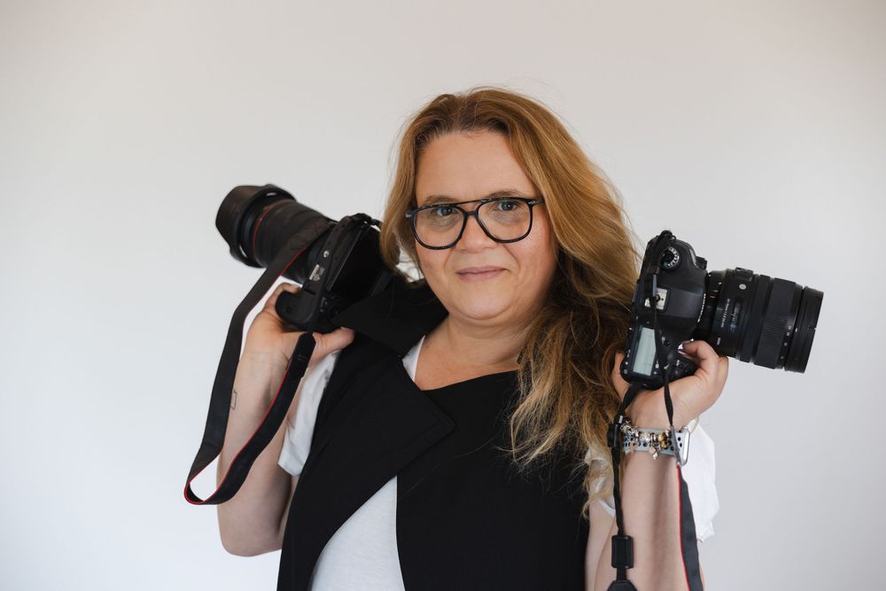 Professional photographer wearing glasses holds camera equipment against white studio background.