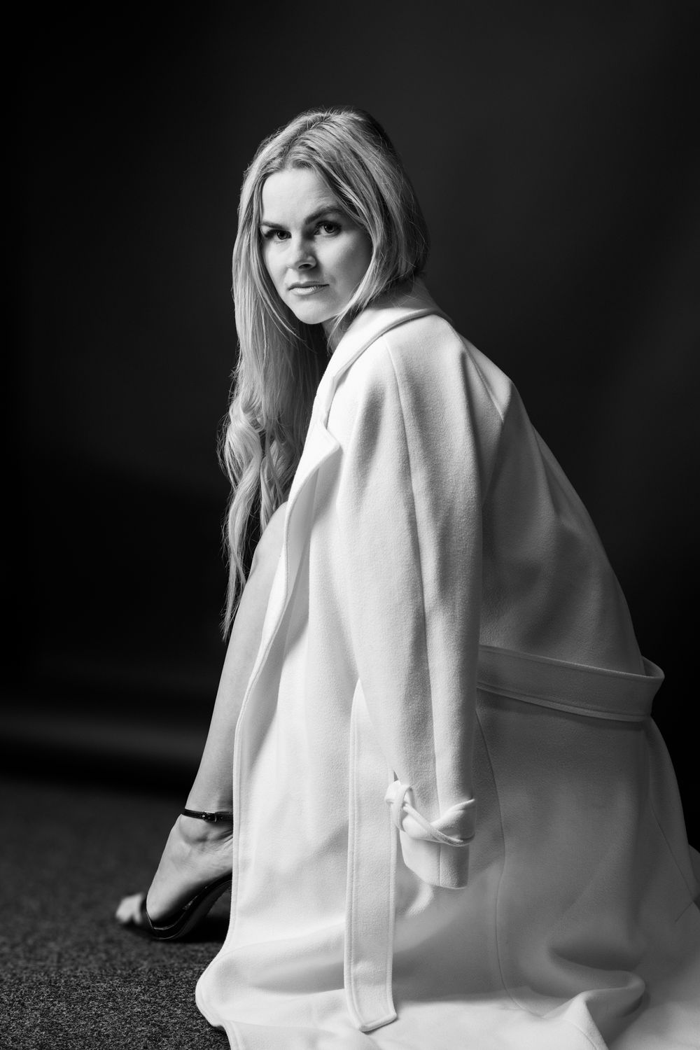 Black and white portrait of a blonde woman in a white coat, looking over her shoulder against a dark background.