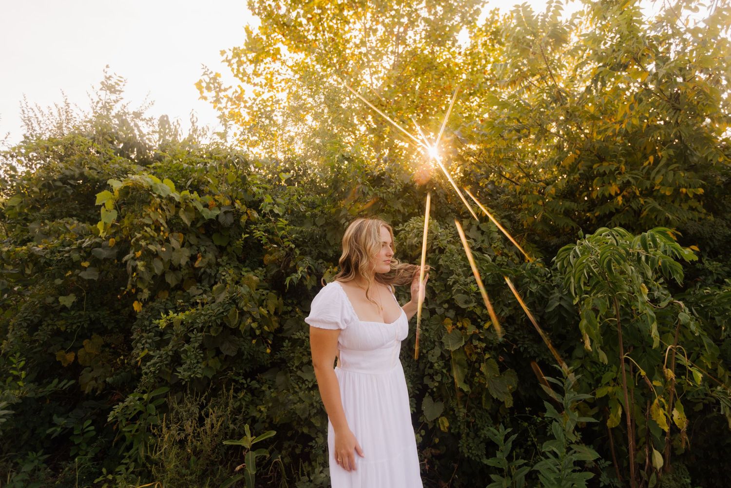 Figure in a white dress holds a glowing wand staff against golden sunset light in a lush garden setting.