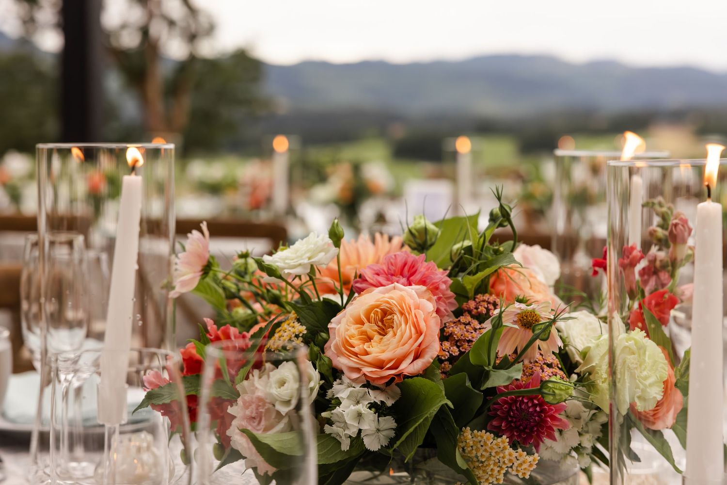 Elegant wedding table centerpiece with peach roses, greenery and glowing candles against a mountain landscape backdrop.