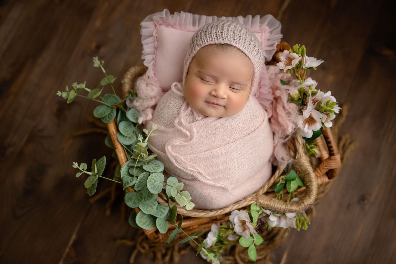 A sleeping newborn wrapped in soft pink swaddle lies in a rustic wooden basket with delicate flowers and greenery.