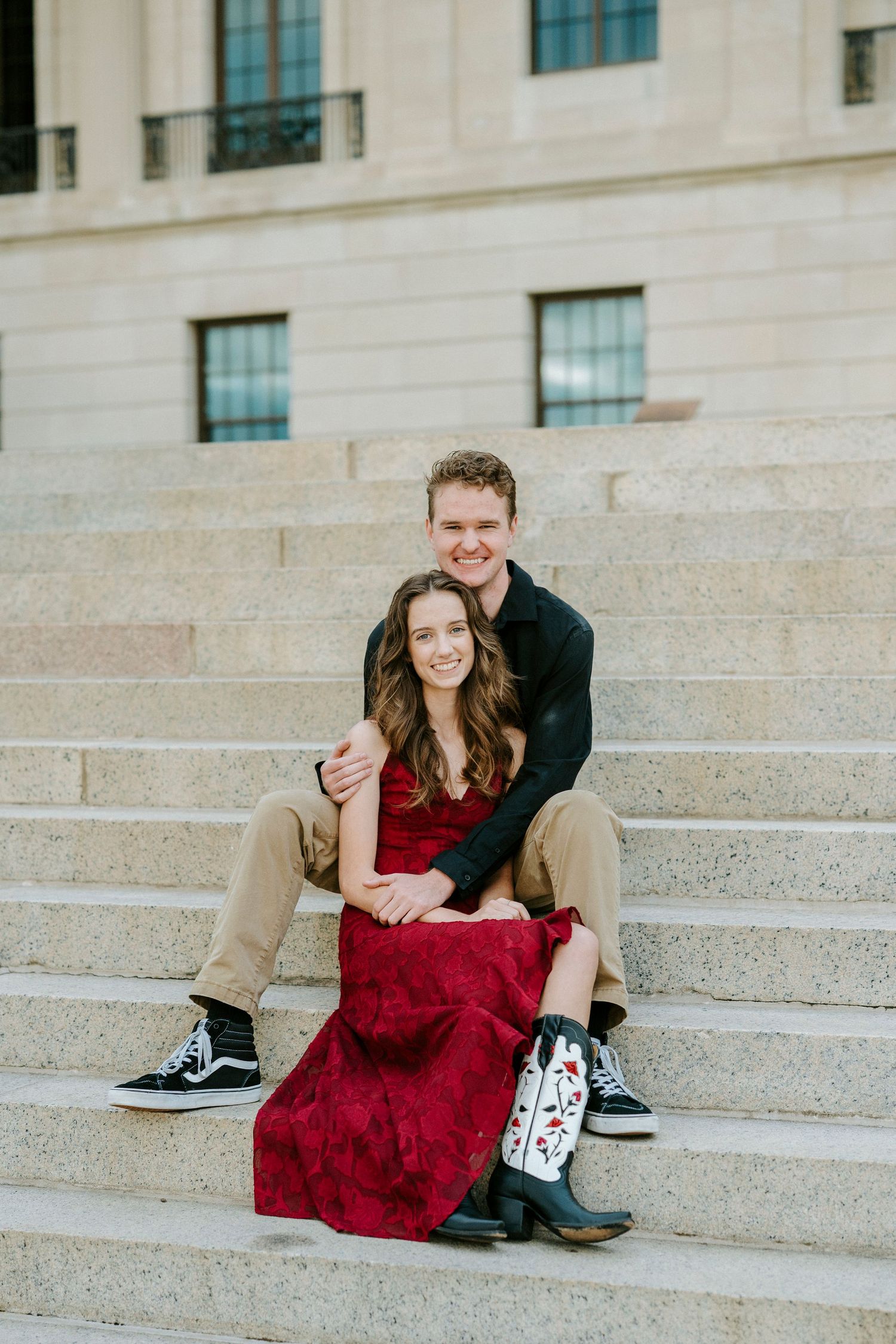 A couple sits together on concrete steps outside a building, with the person in a red dress and boots.