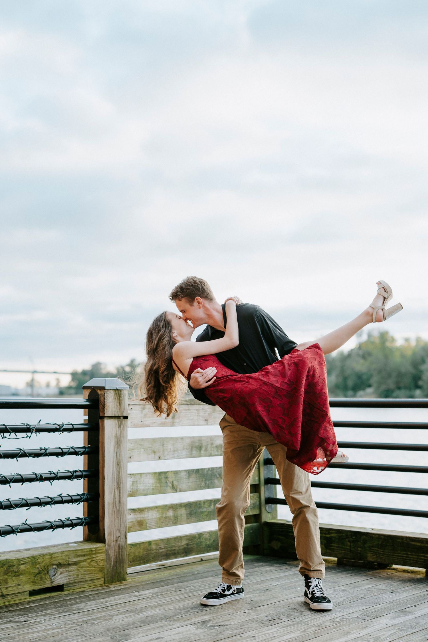 A romantic couple in a dip pose on a wooden deck overlooking a scenic landscape on a cloudy day.