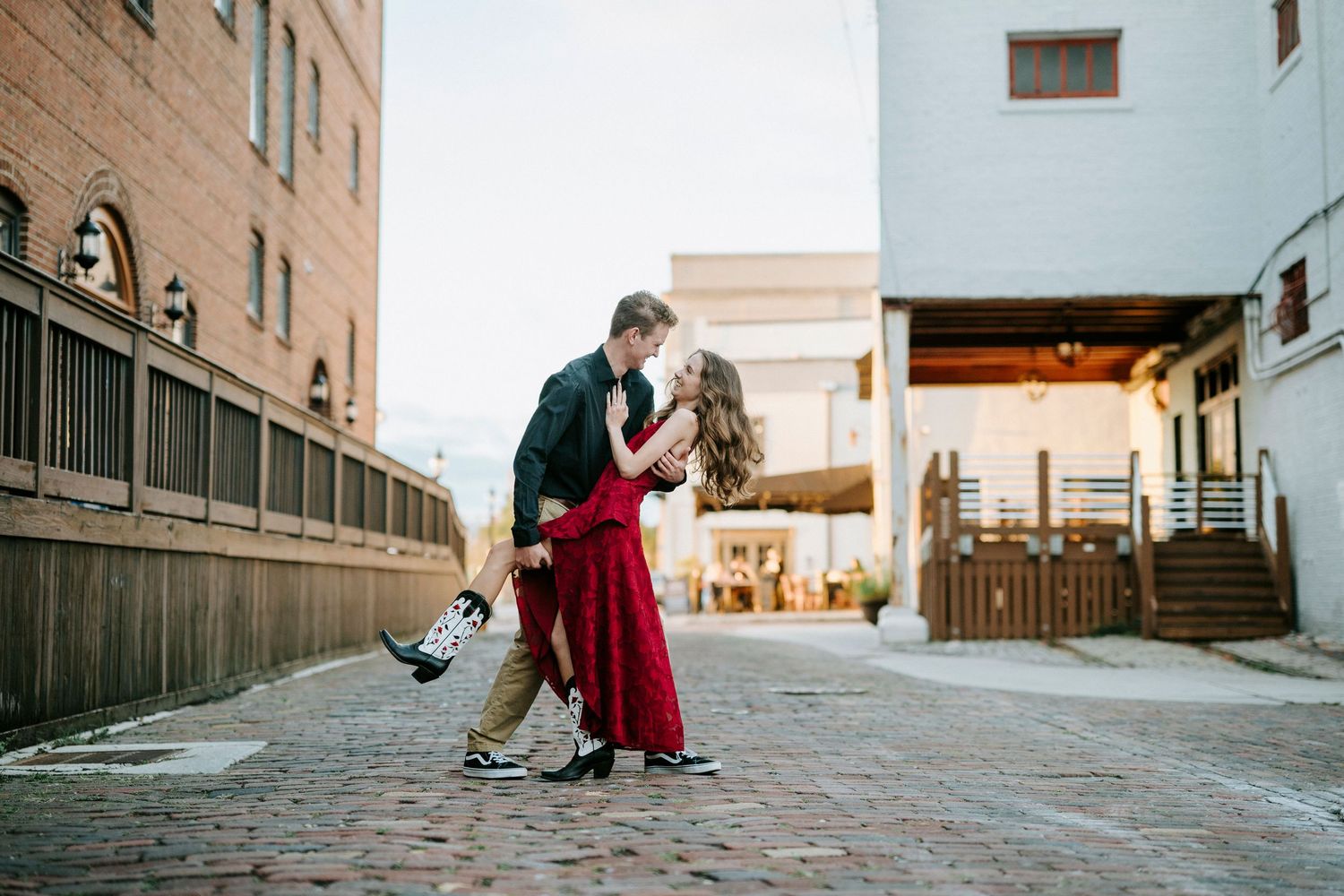 A couple shares an intimate moment on a cobblestone street beside brick buildings in an urban setting.