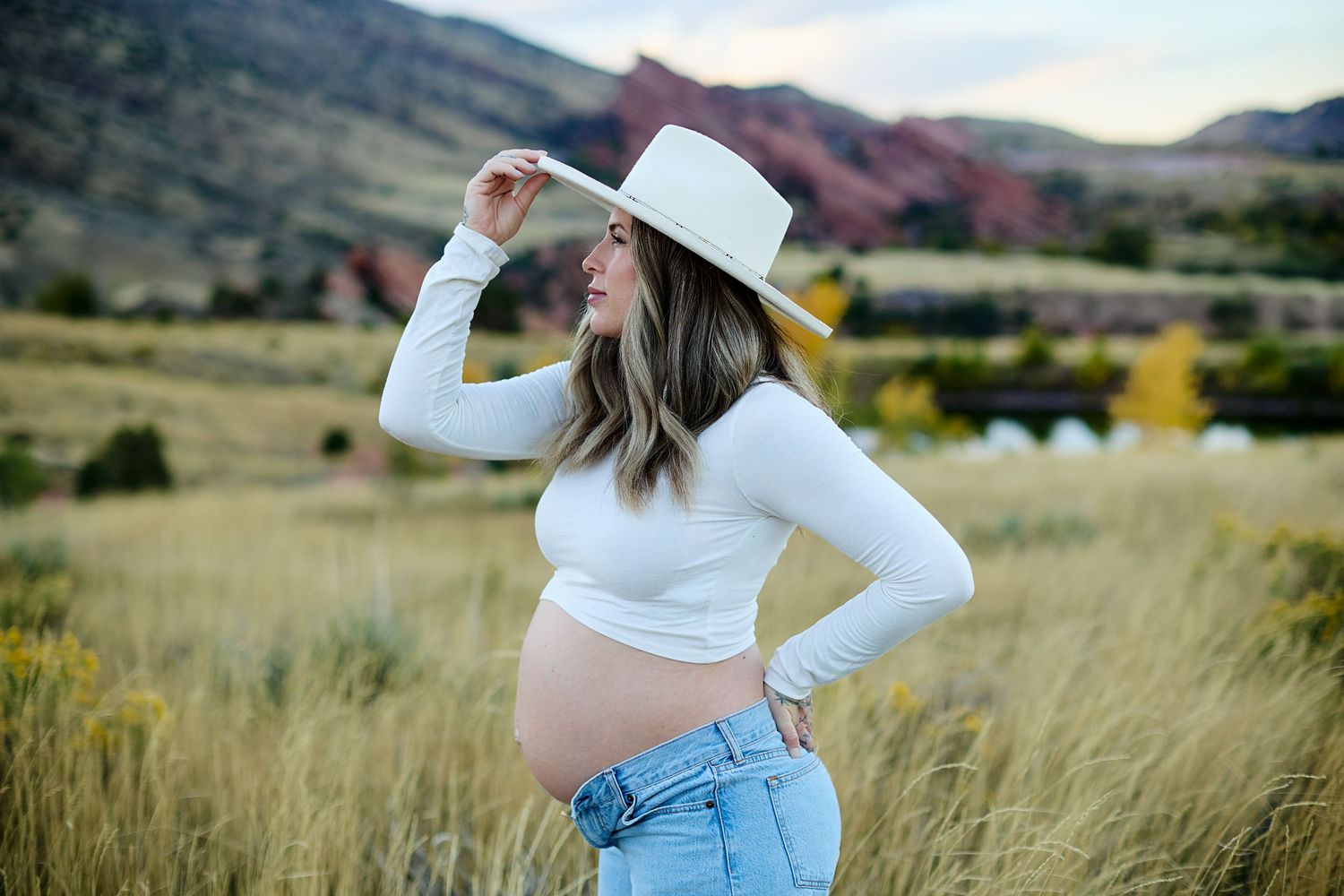 A person in a white top and cowboy hat poses gracefully in a golden field with mountains in the background.