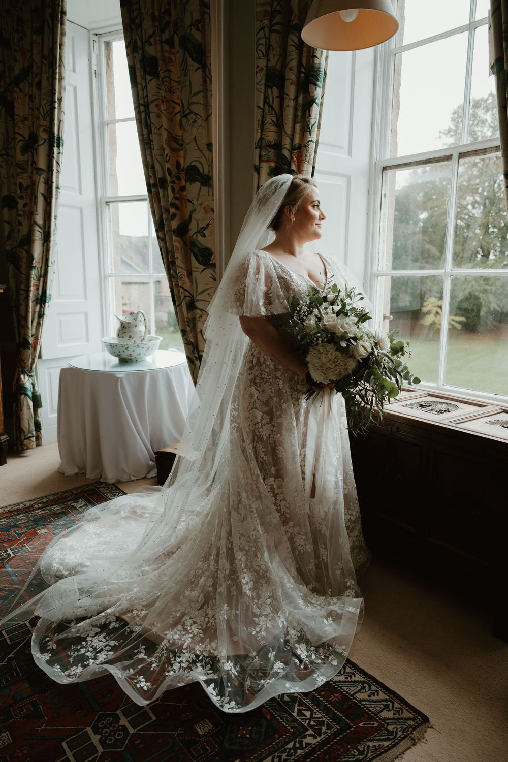 Bride in lace wedding gown poses elegantly by sunlit window with white bouquet in vintage-style interior.