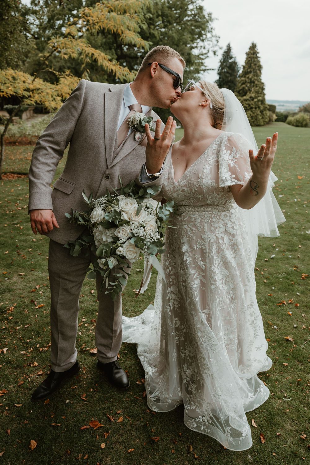 Romantic outdoor wedding scene with couple embracing on grass with autumn trees in background.