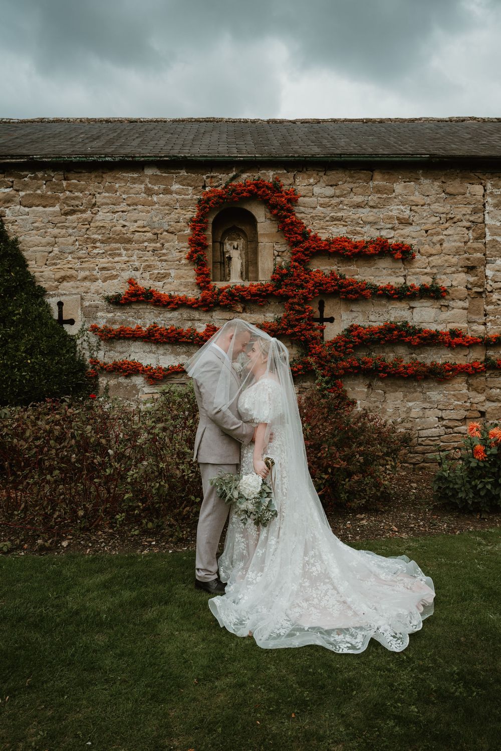 A romantic wedding moment captured against a rustic stone wall adorned with vibrant red climbing flowers.