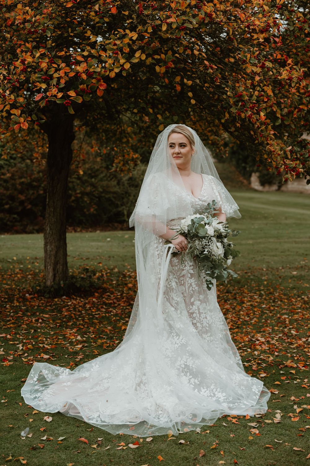 A bride in a flowing lace wedding gown sits gracefully among autumn leaves beneath trees with orange foliage.