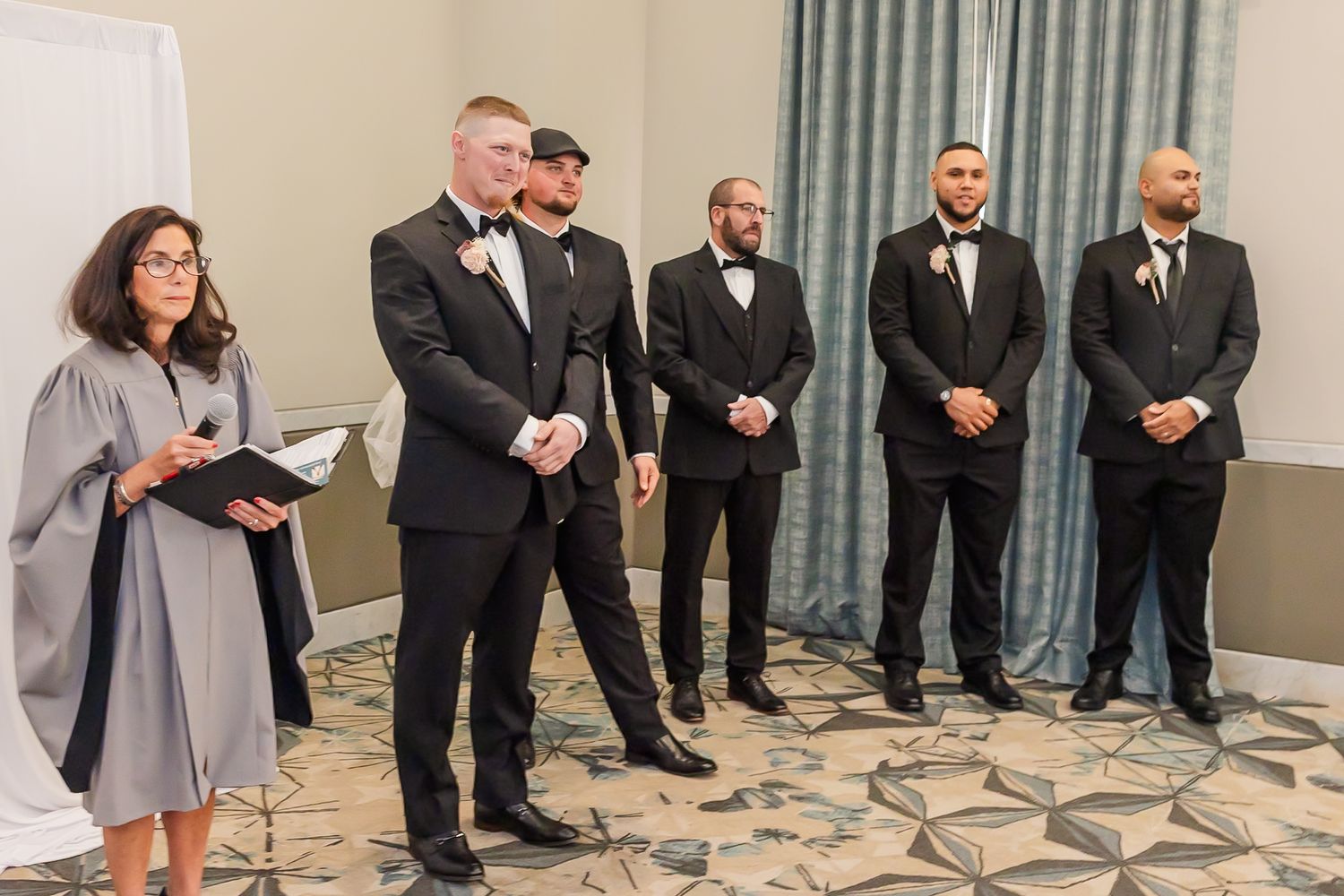 Wedding party members in black tuxedos and gray dresses stand in a line against a light-colored wall.