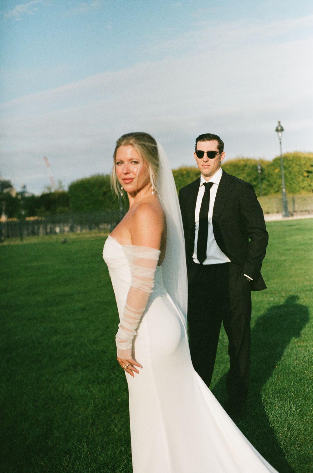 A wedding couple poses together on a grassy lawn during a sunny outdoor photo session.