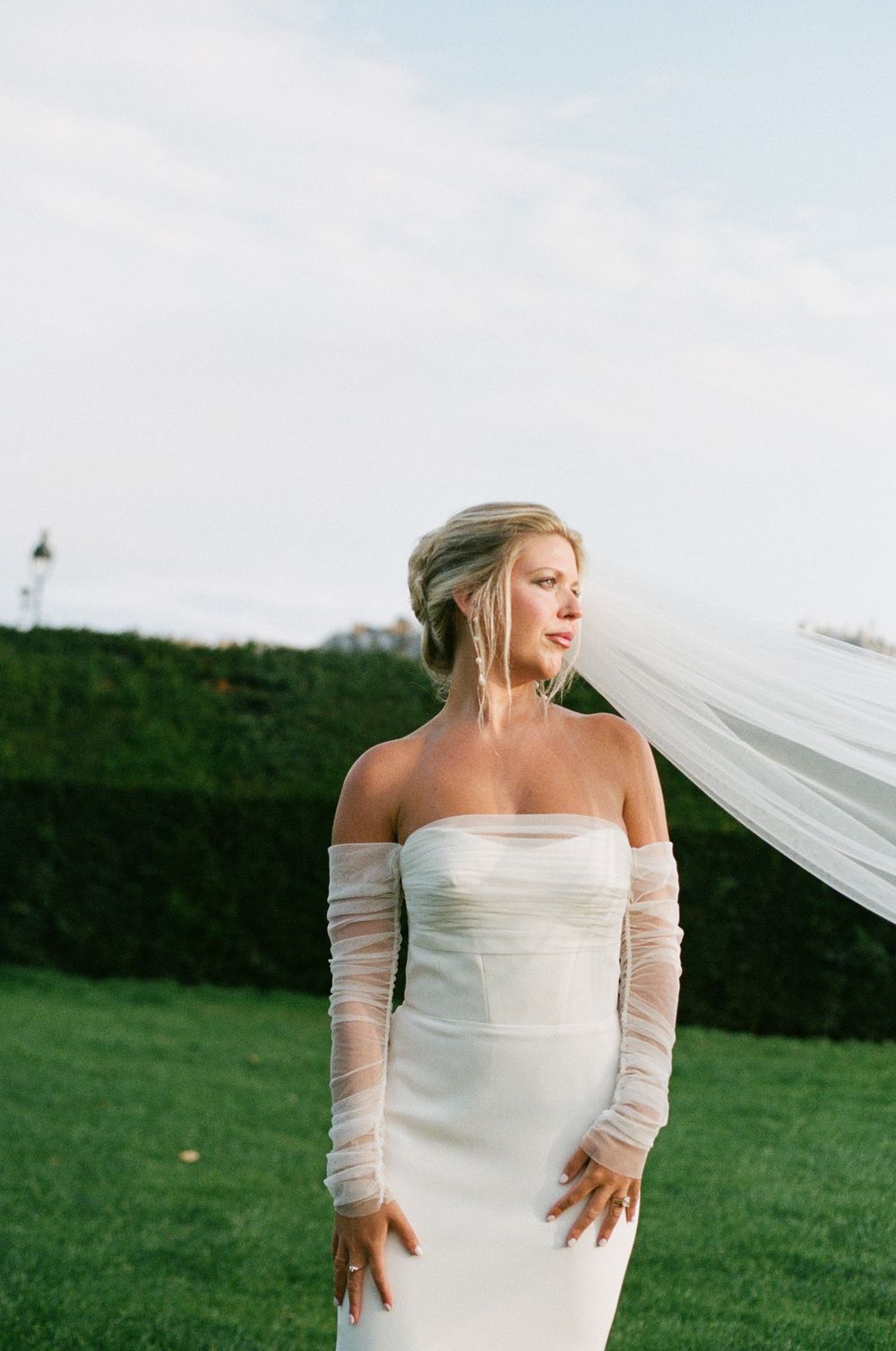 A bride in an elegant white off-shoulder wedding dress poses playfully with her veil flowing in the wind.