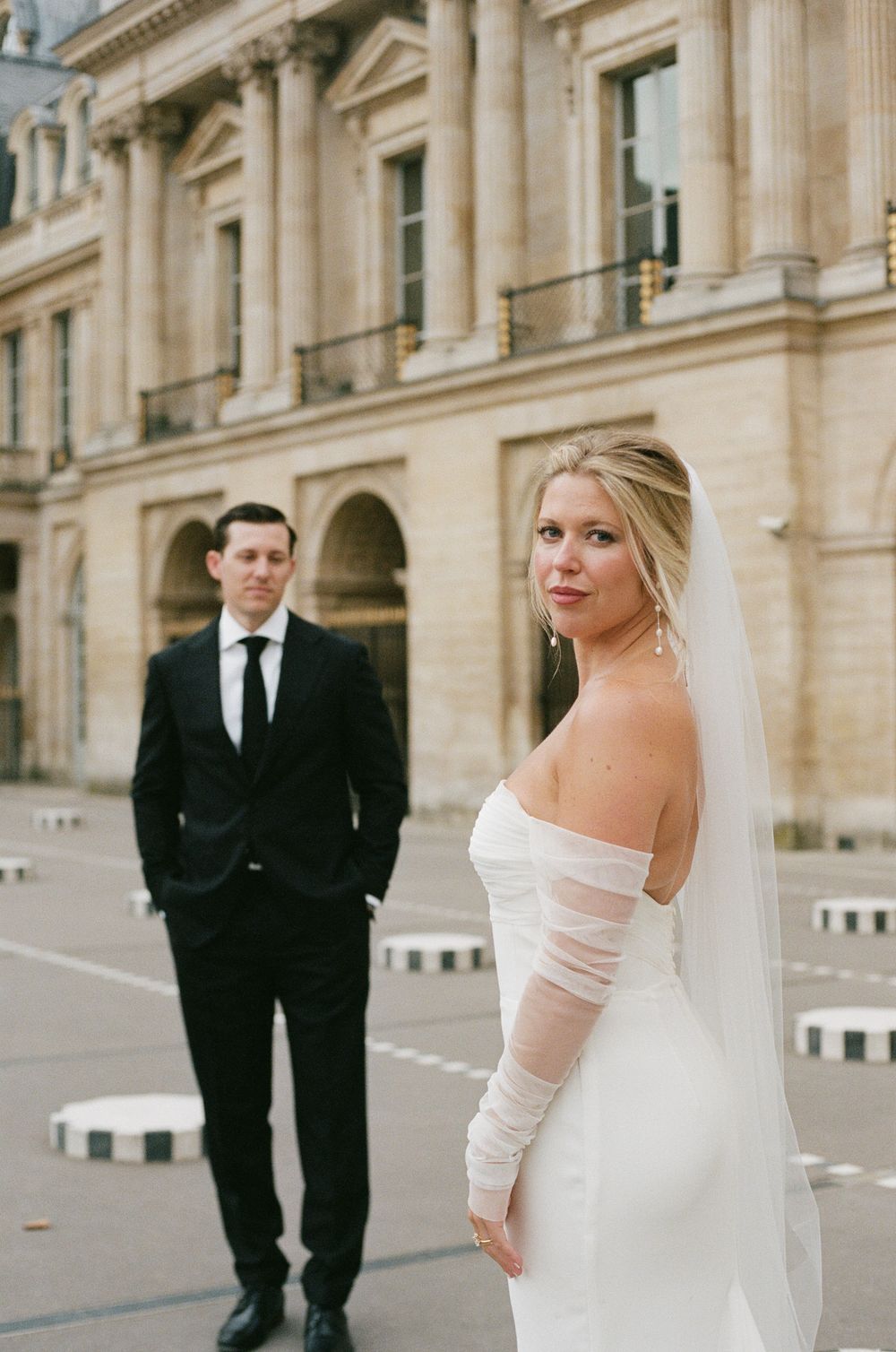 A wedding couple poses in front of a grand stone building with classical architecture and columns. Paris editorial elopement photographer.