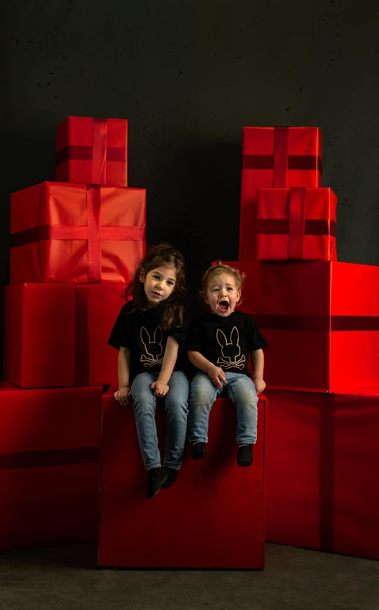 Two children sit and laugh on red blocks against a dark background in a creative studio setting.