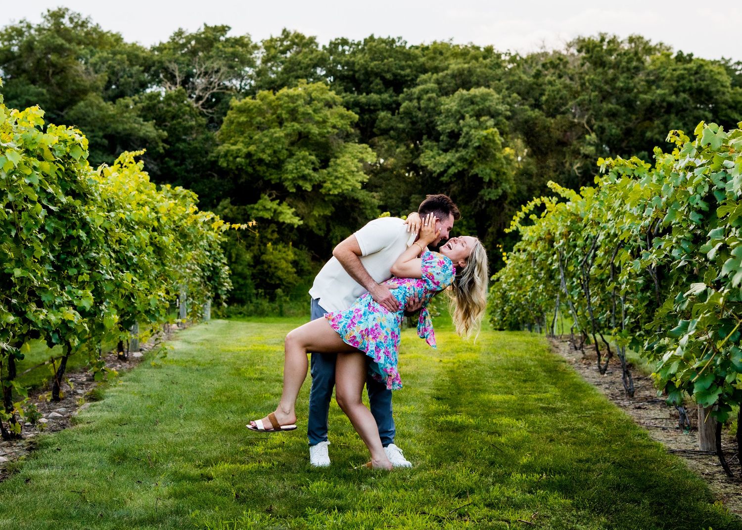 A couple embraces romantically in a lush vineyard at sunset with rows of grape vines stretching into the distance.