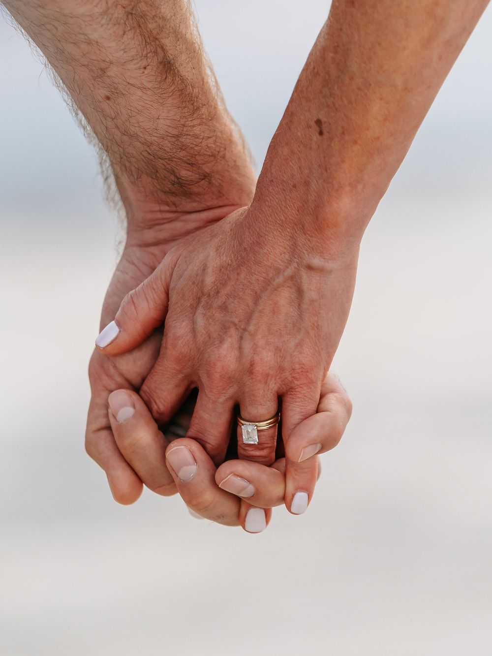 Close up of two hands clasped together showing a wedding ring against blurred beach background.