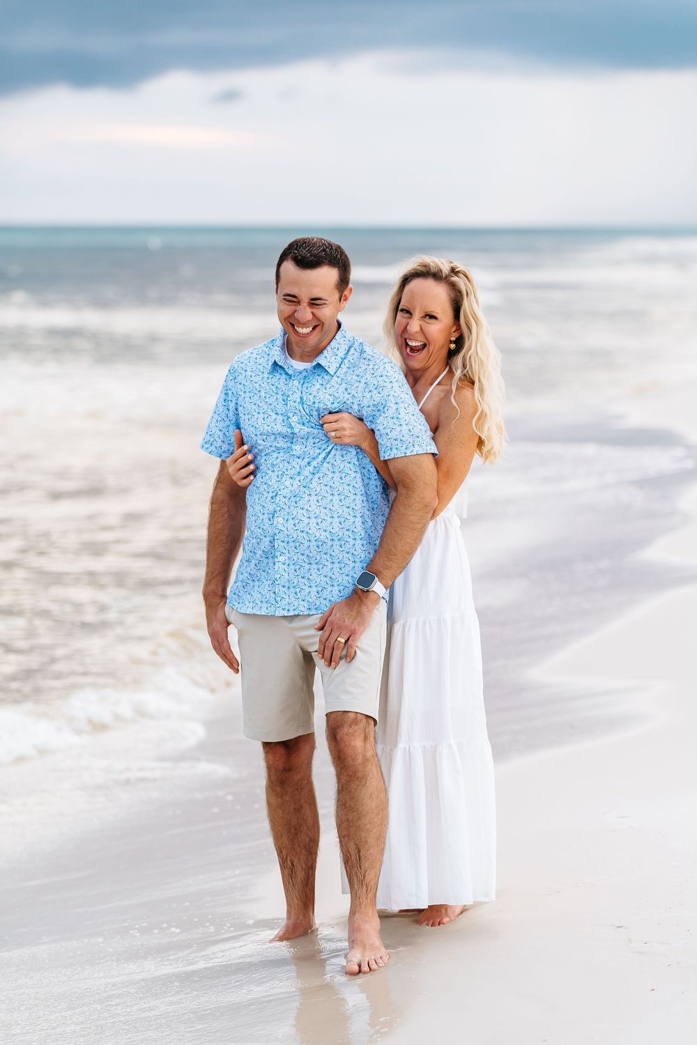 Playful couple walking and laughing along shoreline with dramatic storm clouds over ocean.