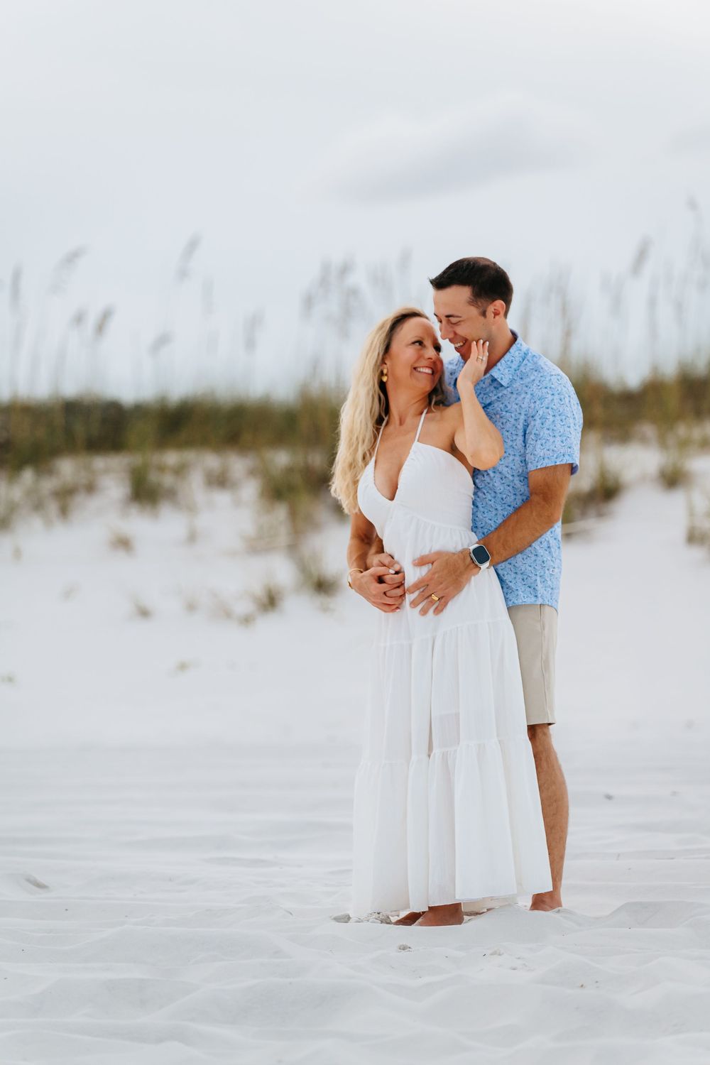 A couple shares intimate moments on a pristine white sand beach with sea oats and dunes in the background.