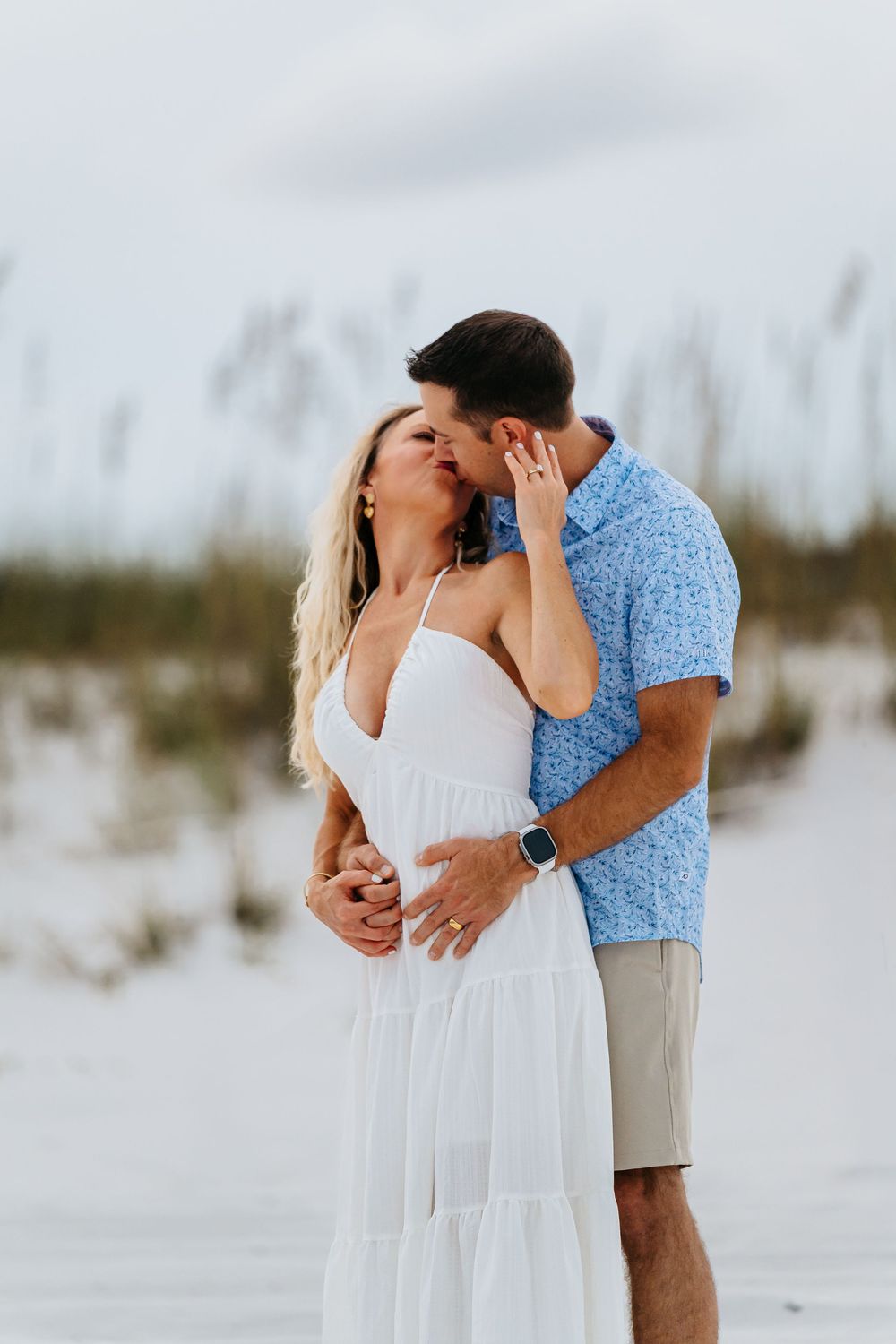 A romantic couple embraces on a white sandy beach with sea oats swaying in the background.