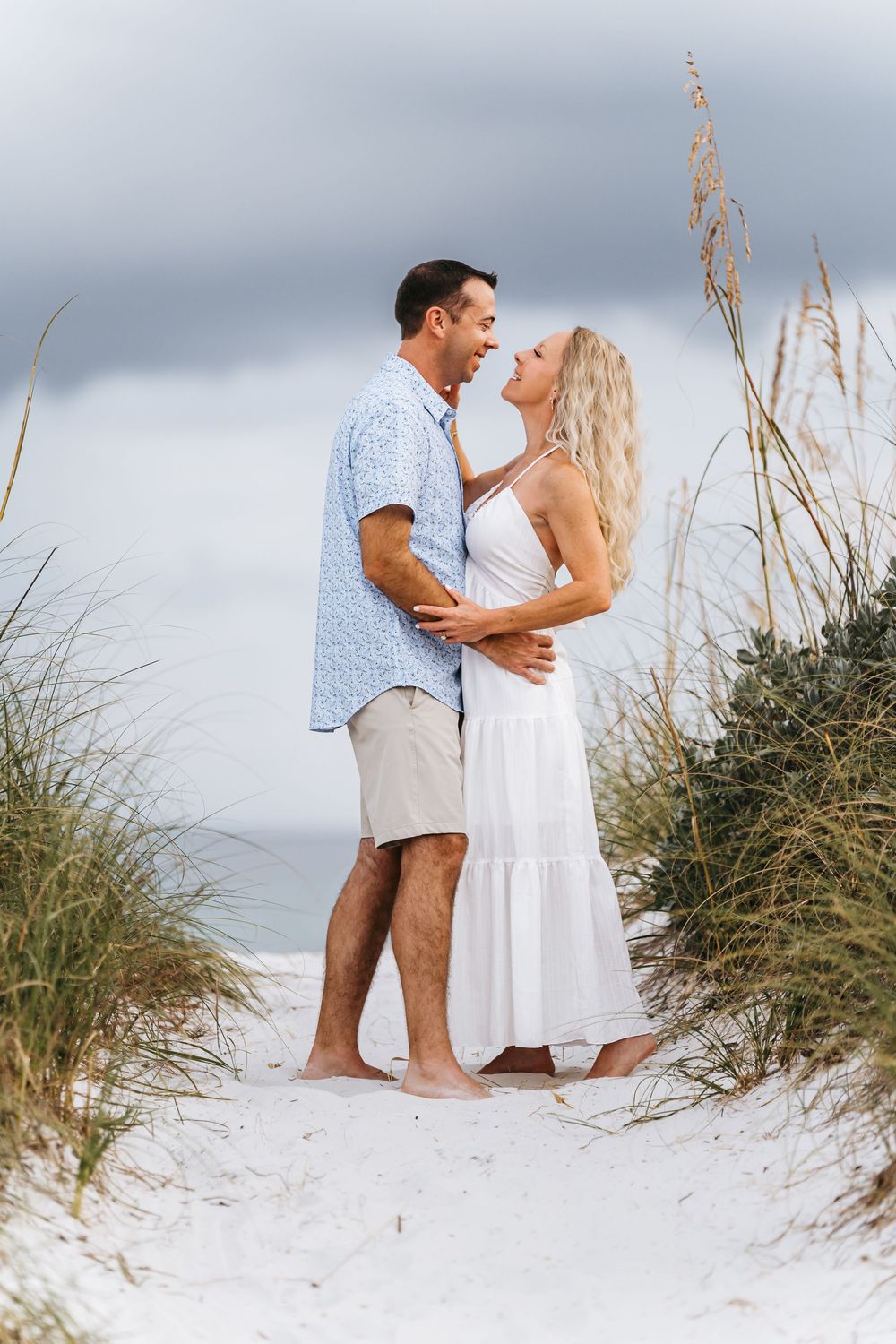 A couple in white attire shares a romantic moment on a sandy beach with sea grass in the background.