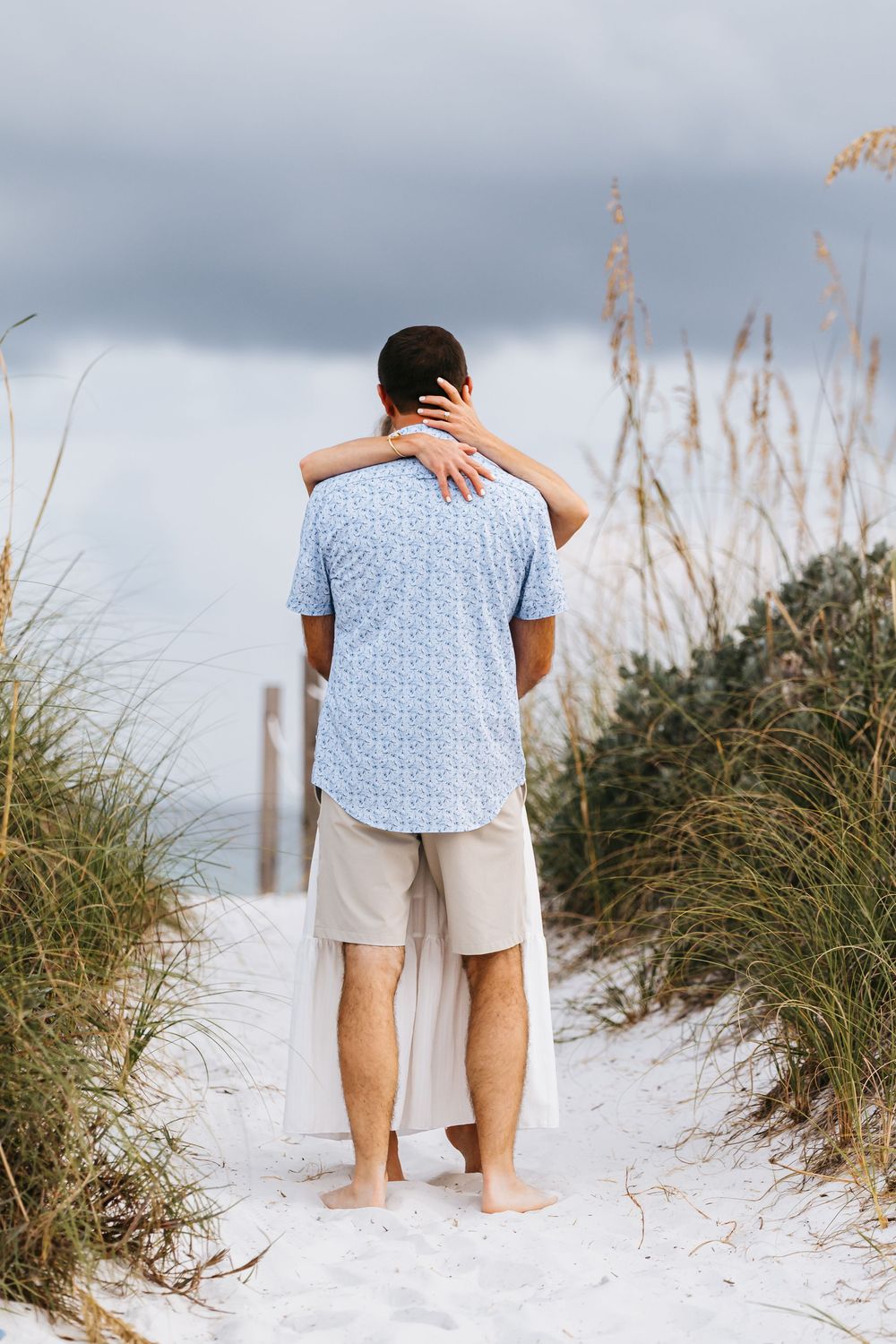 Person standing barefoot on sandy beach path through sea grass on cloudy day wearing blue shirt and khaki shorts.