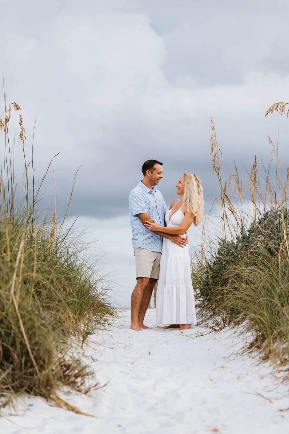 A couple shares a romantic moment on a white sandy beach with sea oats and cloudy skies in the background.
