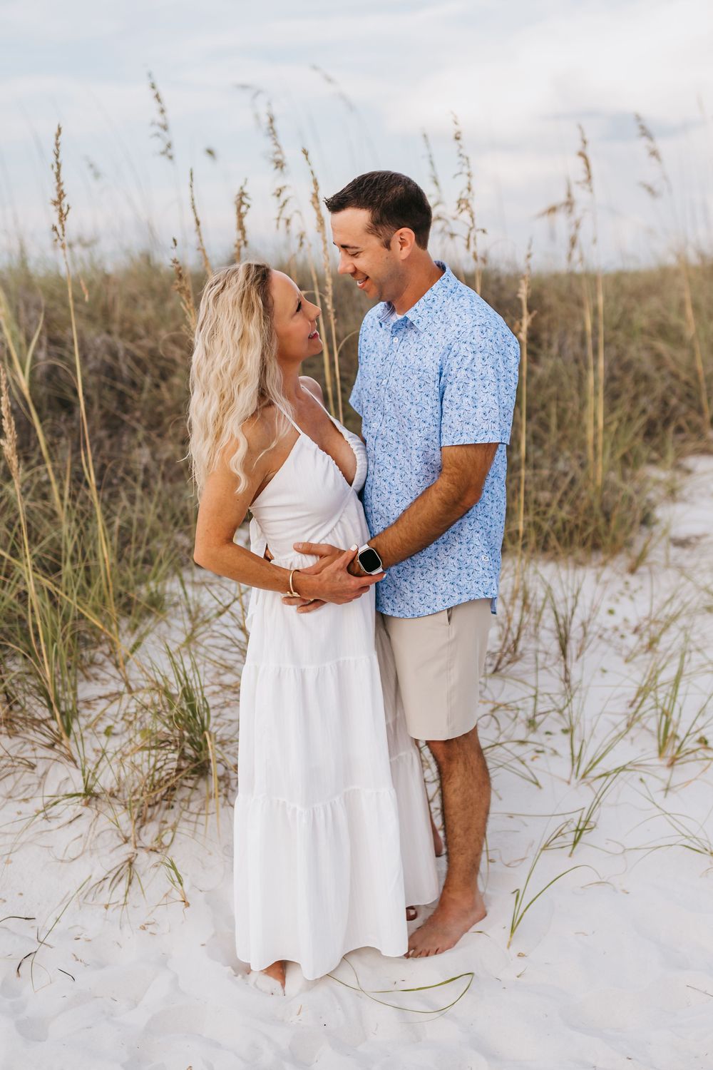 Couple embracing on white sandy beach with sea oats swaying in the background on a sunny day.