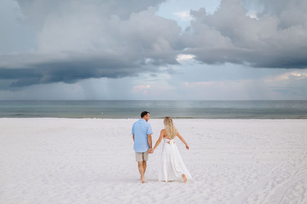 Couple walking hand in hand on pristine white sandy beach with stormy clouds overhead at sunset.