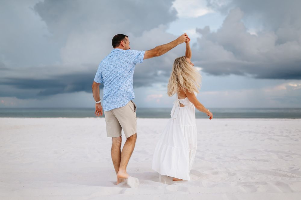 Couple in white and blue outfits walking and dancing together on a pristine white sandy beach under cloudy skies.