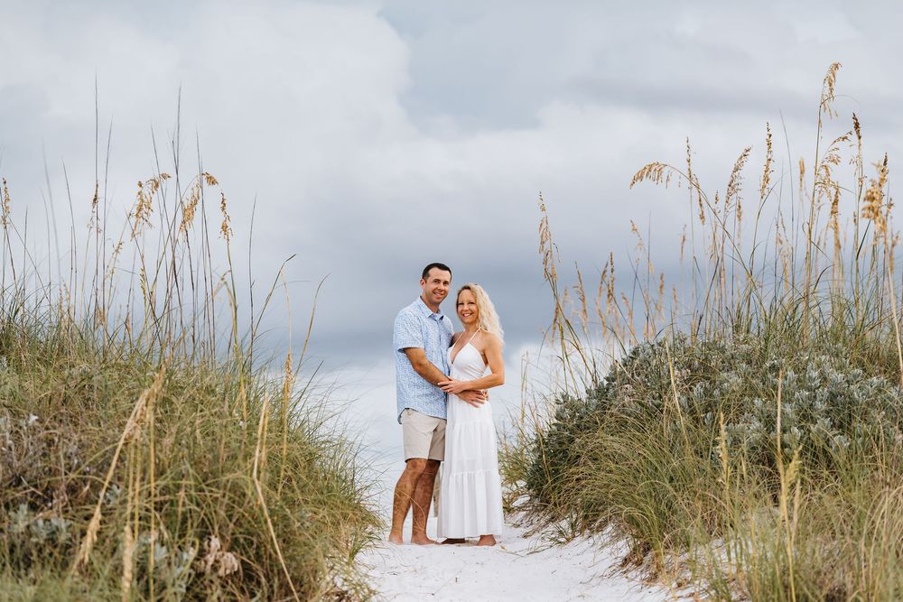 A couple stands together on a sandy beach path surrounded by tall sea grass and dunes on a cloudy day.