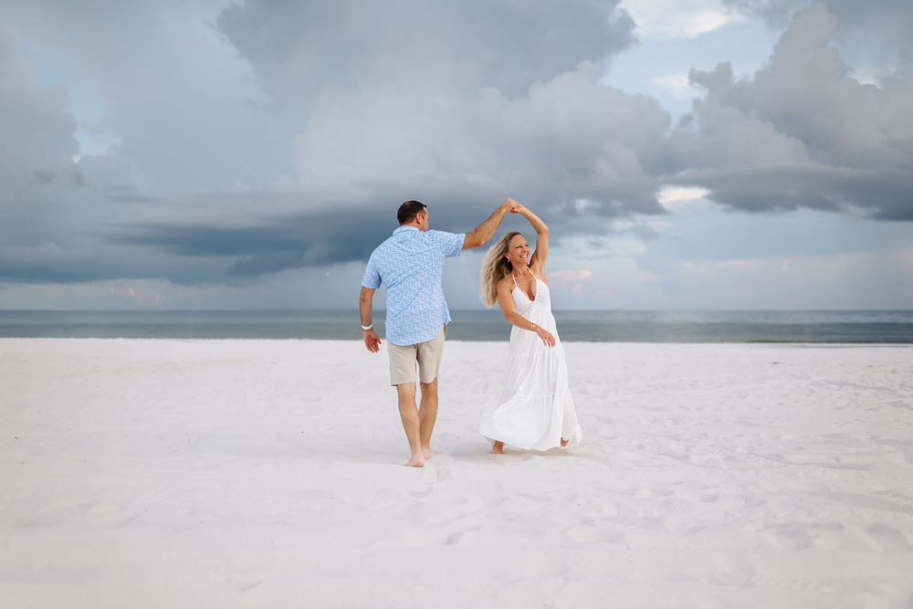 Two people walk along a pristine white sand beach with dramatic cloudy skies and ocean views.