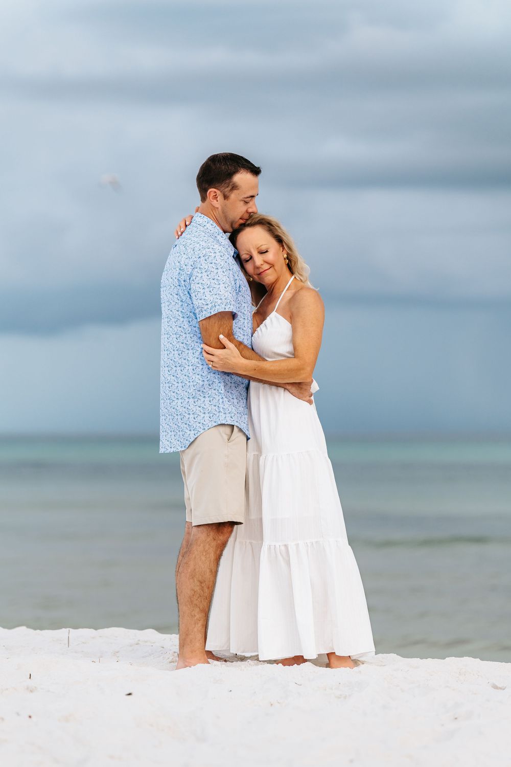 A couple embraces tenderly on a white sandy beach with the ocean and cloudy sky behind them.
