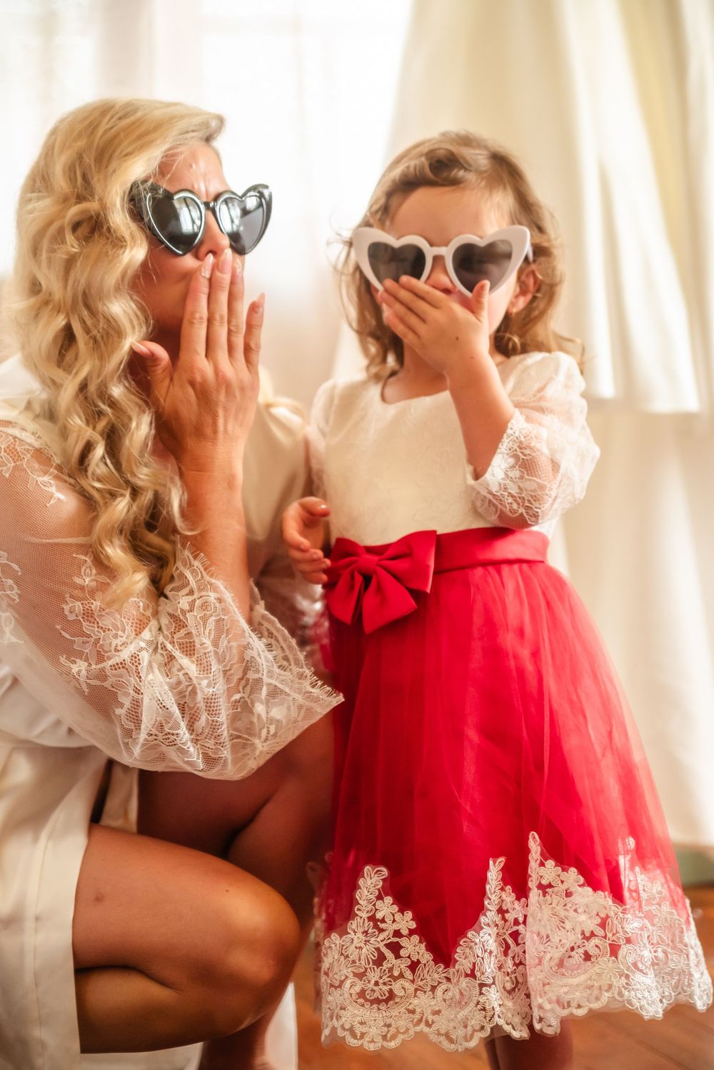 Two people in red and white lace dresses share an intimate moment in front of sunlit curtains.