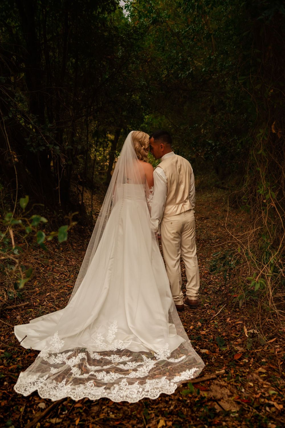 Wedding couple in white attire walking through autumn leaves with lace train trailing behind.