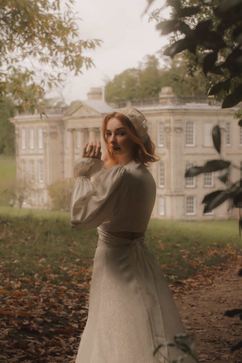 A person in a light coat spins playfully on autumn leaves in front of a grand historic manor house.