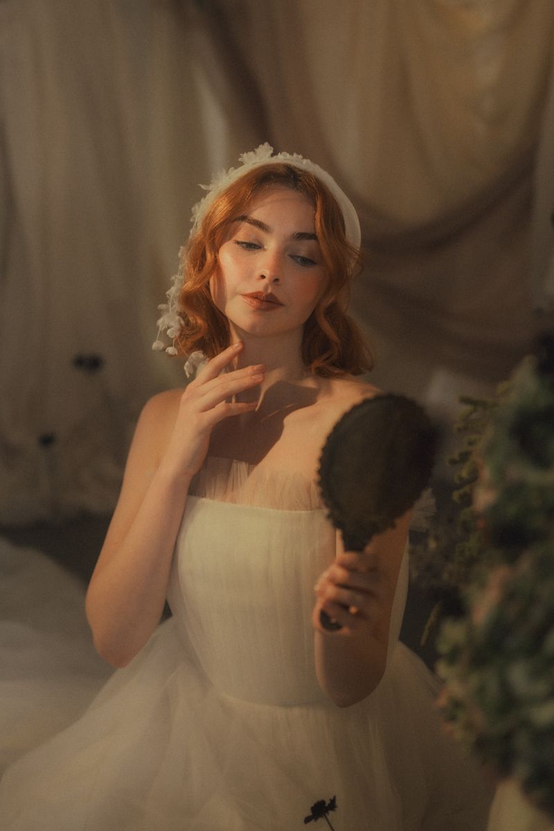 A dreamy portrait in soft lighting shows a person in a white tulle dress holding a vintage hand mirror.