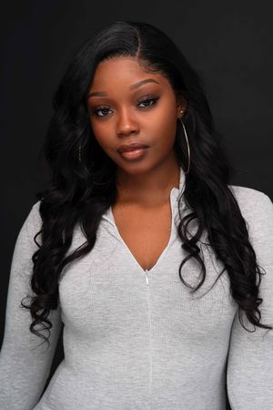 Black woman with long wavy hair in gray zip-up top poses against dark background.