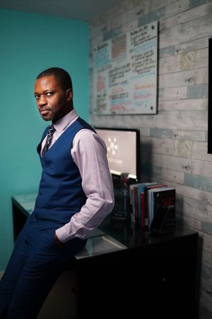 Black man in blue vest and tie leans against desk in modern office.