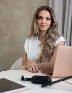 Young blonde woman in white t-shirt sits at desk with laptop smiling.