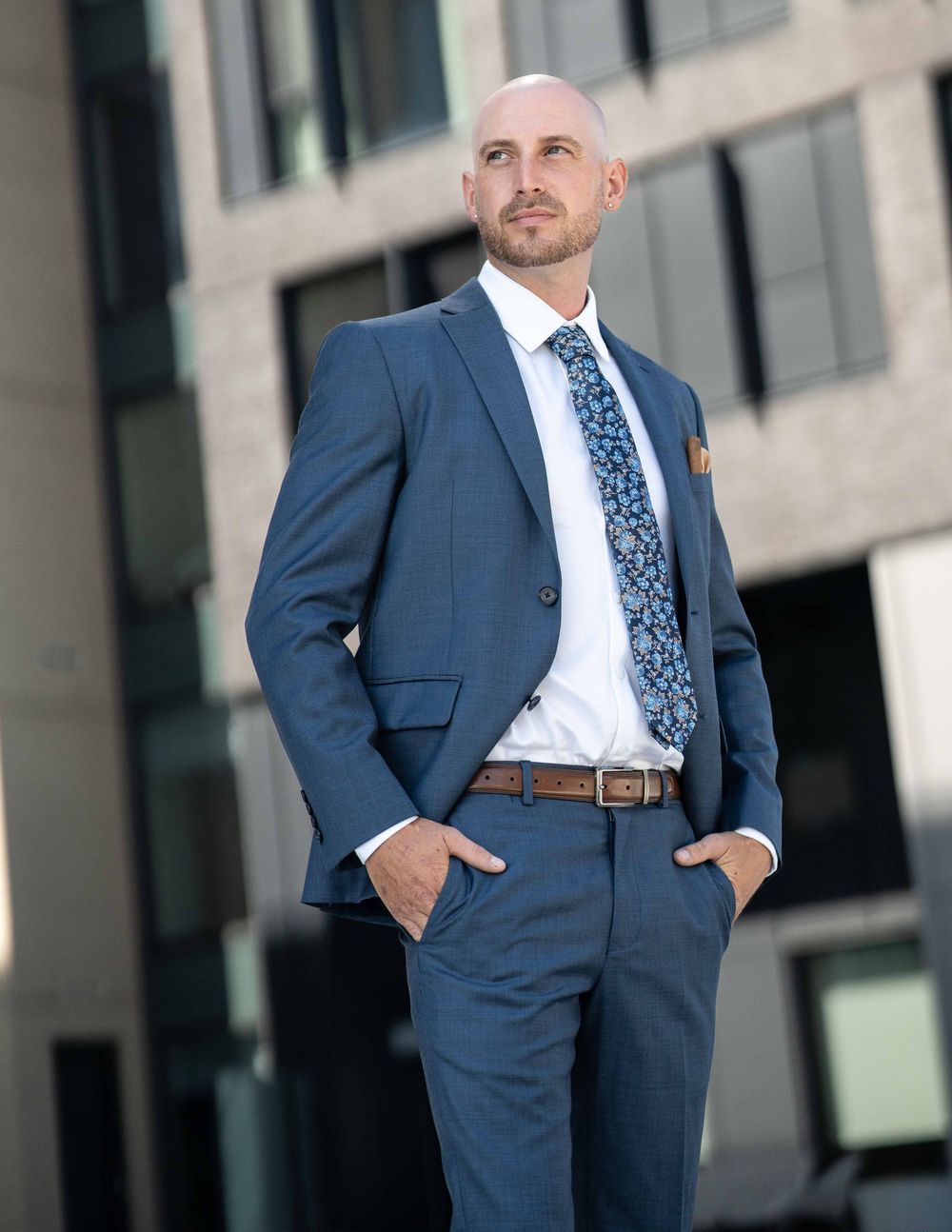 Bald professional man in navy blue suit poses outdoors near modern building.