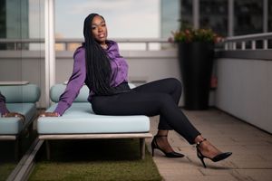 Black woman in purple blouse and black pants poses on rooftop terrace at dusk.