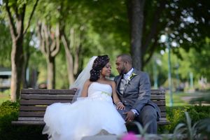 Bride and groom sit on park bench gazing at each other surrounded by lush green trees.