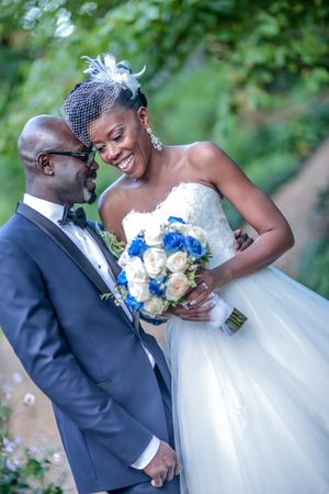 Smiling bride in strapless lace gown holds blue and white bouquet beside groom in tuxedo outdoors.